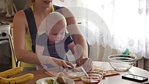 Young woman with a toddler boy sitting in a sling cuts eggs while sitting at the kitchen table and, after finishing