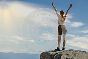 The young woman at the top of the mountain raised her hands up on blue sky background. The woman climbed to the top and enjoyed