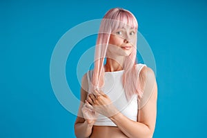 Young woman touching her smooth natural long pink dyed hair and looking surprised, posing isolated over blue studio