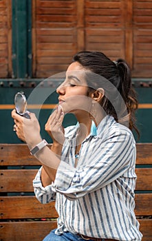 Young woman wearing casual clothes putting on makeup sitting on a park bench