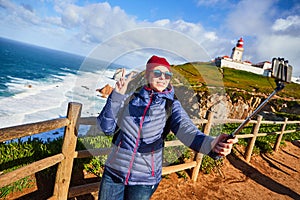 Young woman tourist at Cape Caba da Roca take a selfie shoots with view on beautiful landscape on a mobile phone, winter trip to photo