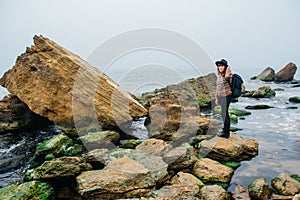 Young woman tourist in hat and with backpack standing on a rock against a beautiful sea, looking at sea, on horizon. Place for