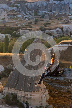 A young woman tourist in a hat sits on a mountain and looks at the sunrise in Cappadocia. Turkey. Top attraction travel destinatio