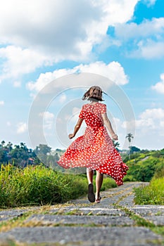 Young woman tourist in a lon red dress running on the rainforest trail. Bali island.