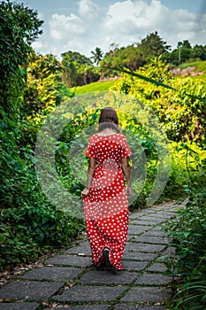 Young woman tourist in a lon red dress walking on the rainforest trail. Bali island.
