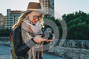 Young woman tourist, photographer, hipster girl in hat stands on city street and uses camera, looks images on screen.