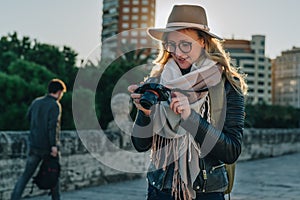 Young woman tourist, photographer, hipster girl in hat stands on city street and uses camera, looks images on screen.