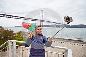 Young woman tourist take selfies on the background of famous iron 25th of April bridge and holding the flag of Portugal in hands