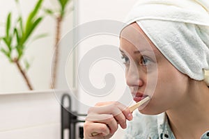 A young woman with a towel on her head in a bright bathroom with a toothbrush in her hand brushes her teeth.