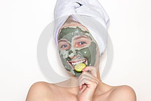 A young woman with a towel on her head and a clay mask on her face and a piece of cucumber. Skin care at home. White background