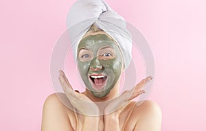 A young woman with a towel on her head and a clay mask on her face on a pink background. Face care, Spa treatments at home