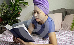 a young woman with a towel on her head is lying in bed and reading a book. The concept of rest, education and time for