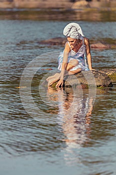 Young woman with a towel on her head at the sea. The lady touches the water with her hands. The concept of summer outdoor recreati
