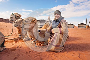 Young woman in traditional Bedouin coat - bisht - and headscarf crouching next to two camels laying on red desert ground, smiling photo