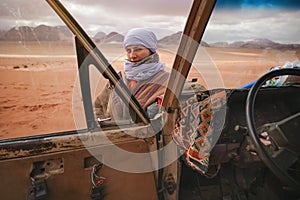 Young woman in traditional Bedouin coat - bisht - and headscarf, posing next to old 4wd vehicle, looking over opened door - desert photo