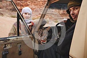 Young woman in traditional Bedouin coat - bisht - and headscarf, sitting next to old 4wd vehicle, looking over opened door, photo