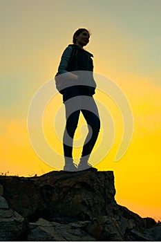 Young Woman Traveler with Backpack Standing on the Top of the Rock at Summer Sunset. Travel and Adventure Concept.