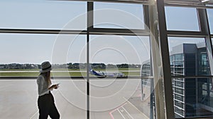 Young woman traveler, leisure or business standing at the glass window of a large international airport hub with a phone