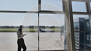 Young woman traveler, leisure or business standing at the glass window of a large international airport hub with a phone