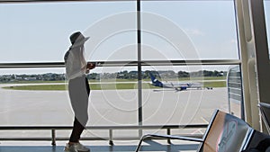 Young woman traveler, leisure or business standing at the glass window of a large international airport hub with a phone