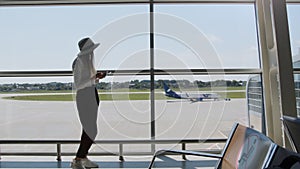 Young woman traveler, leisure or business standing at the glass window of a large international airport hub with a phone