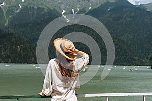 Young woman traveler with long blonde hair in straw hat looks at beautiful view of the mountain lake, people from behin