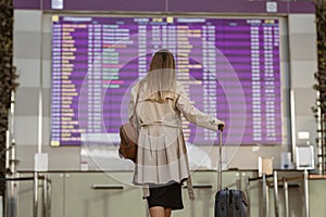 Young woman traveler with luggage looks at information board at airport in search of his flight