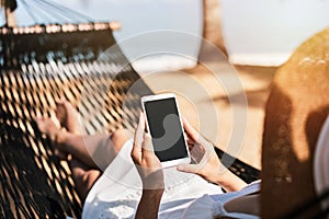 Young woman traveler lying on a hammock and using smartphone at the beach while traveling for summer