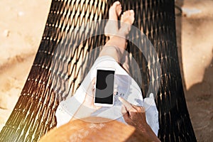 Young woman traveler lying on a hammock and using smartphone at the beach while traveling for summer vacation