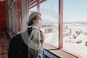 Young woman traveler in a protective medical mask with a backpack looks out the window at the airfield at the airport waiting for