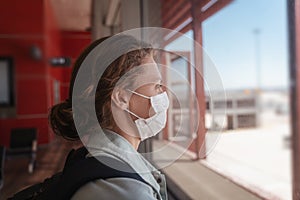 Young woman traveler in a protective medical mask with a backpack looks out the window at the airfield at the airport waiting for