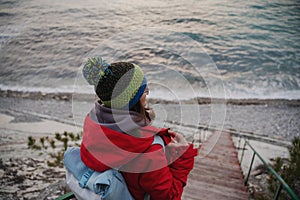 Young woman traveler in a red jacket and hat standing on a hill and looking at the sea. Winter sea travel concept