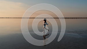 A young woman traveler in a shirt runs along a salt lake with a reflection of the sky against a sunset background