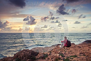 Young woman traveler sits on the shore of the winter sea and watches the bright colorful sunset