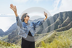 Young woman traveler taking a selfie using a smartphone on the background of the ocean and mountains