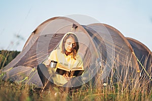 Young woman traveler in yellow hoodie reading book against the tent. Local tourism, weekend trip