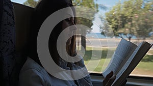 Young woman traveling by bus and reading book. Girl is traveling in the car in front of the window