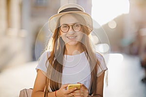 Young woman travels in hat and eyeglasses stands on city street and Texting SMS on her Mobile Phone. Attractive young