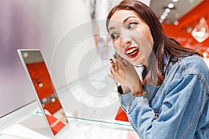 Young woman trying on earrings and looking at the mirror in jewelry store, shopping concept