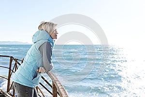 A young woman in a turquoise jacket stands and looks at the sea in the off-season on a sunny day. Cute blonde. Rest and relaxation