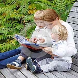 Young woman and two little sibling boys sitting on bench in park and reading fairytale book together. Family having fun and enjoy