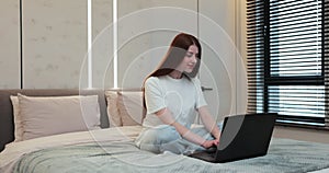 Young woman typing on laptop. Girl typing messages on laptop chatting with friends sitting in bed at home. Female person