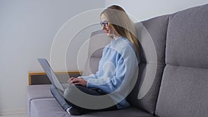 Young woman typing text on laptop keyboard on sofa. Beautiful blonde female sitting on couch at home and coding on notebook pc.