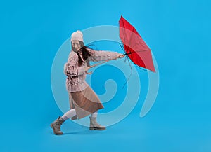 Young woman with umbrella caught in gust of wind on light blue background