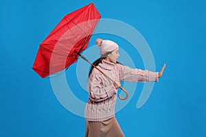 Young woman with umbrella caught in gust of wind on light blue background