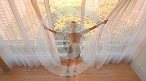 Young woman uncover the big window and looking out her apartment on the city buildings. Sunrise in the city.