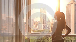 Young woman uncover the big window and looking out her apartment on the city buildings. Sunrise in the city.