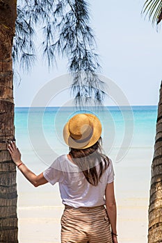A young woman under a palm tree on the shore of the Gulf of Thailand. Woman in a hat looks at the sea
