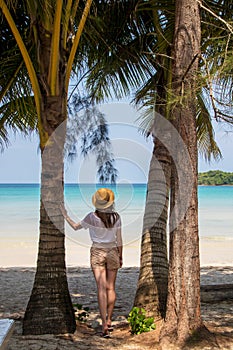 A young woman under a palm tree on the shore of the Gulf of Thailand. Woman in a hat looks at the sea