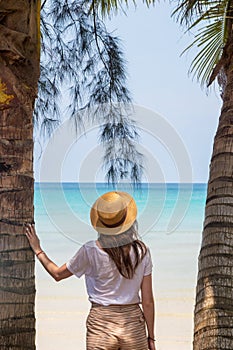 A young woman under a palm tree on the shore of the Gulf of Thailand. Woman in a hat looks at the sea